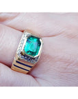 An 18k yellow gold men’s emerald ring with sparkling accent diamonds resting on a finger.