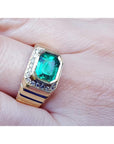 An 18k yellow gold men’s emerald ring with sparkling accent diamonds resting on a finger.