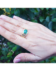 Hand wearing a mens ring with a natural colombian emerald against a green leafy background.