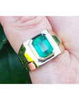 Gold ring with a rectangular emerald-cut on a finger against a blurred green background.