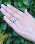 Hand wearing a natural emerald ring against a green leafy background.