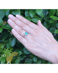 Hand wearing a natural emerald ring against a green leafy background.