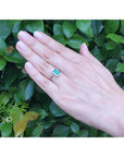Emerald-cut Muzo emerald halo diamond ring in 14K white gold on a hand with green foliage background.