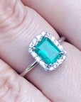 Emerald ring with diamond halo on a finger.