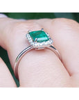 Emerald-cut Muzo emerald halo diamond ring in 14K white gold on a finger.