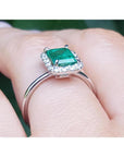 Emerald-cut Muzo emerald halo diamond ring in 14K white gold on a finger.