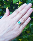 A woman's hand wearing a real emerald ring on a green leafy background.