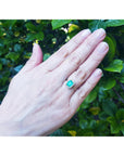 A woman's hand wearing a real emerald ring on a green leafy background.