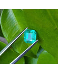 1.55 carat emerald-cut Muzo Colombian loose emerald, sparkling medium bluish green, held by tweezers on a leaf.