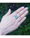 Hand wearing a men's ring with an emerald-cut emerald against a green leafy background.