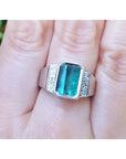 An 18k white gold men’s ring featuring a natural emerald in a bezel setting, accented with diamonds, worn on a finger.