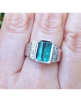 An 18k white gold men’s ring featuring a natural emerald in a bezel setting, accented with diamonds, worn on a finger.