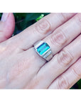 A white gold emerald ring for men on a hand against a blurred green background.
