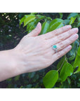Hand wearing a natural emerald ring with accent diamonds against a leafy background.