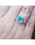 Emerald-cut natural emerald ring with halo diamonds on an 18K white gold split shank band.