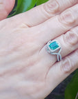 Hand wearing a brides' May birthstone emerald ring against a leafy background.