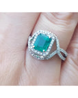 A square emerald ring with halo diamond accents in 18k white gold on a finger.