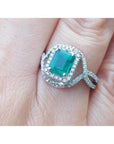 A square emerald ring with halo diamond accents in 18k white gold on a finger.