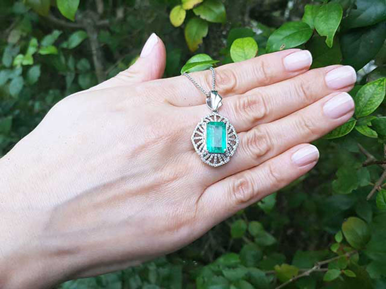 Hand holding a white gold pendant with a rectangular emerald against a green leafy background.