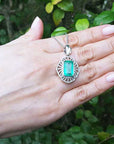 Hand holding a white gold pendant with a rectangular emerald against a green leafy background.