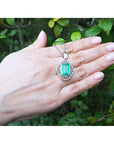Hand holding a white gold pendant with a rectangular emerald against a green leafy background.