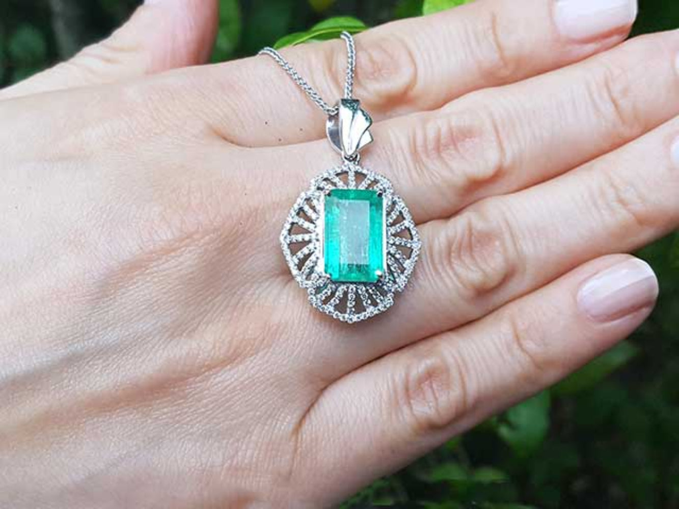 Emerald rectangular gemstone pendant on a white gold chain held in a hand with a blurred green background.