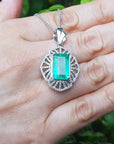 Emerald rectangular gemstone pendant on a white gold chain held in a hand with a blurred green background.