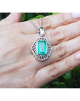 Emerald rectangular gemstone pendant on a white gold chain held in a hand with a blurred green background.