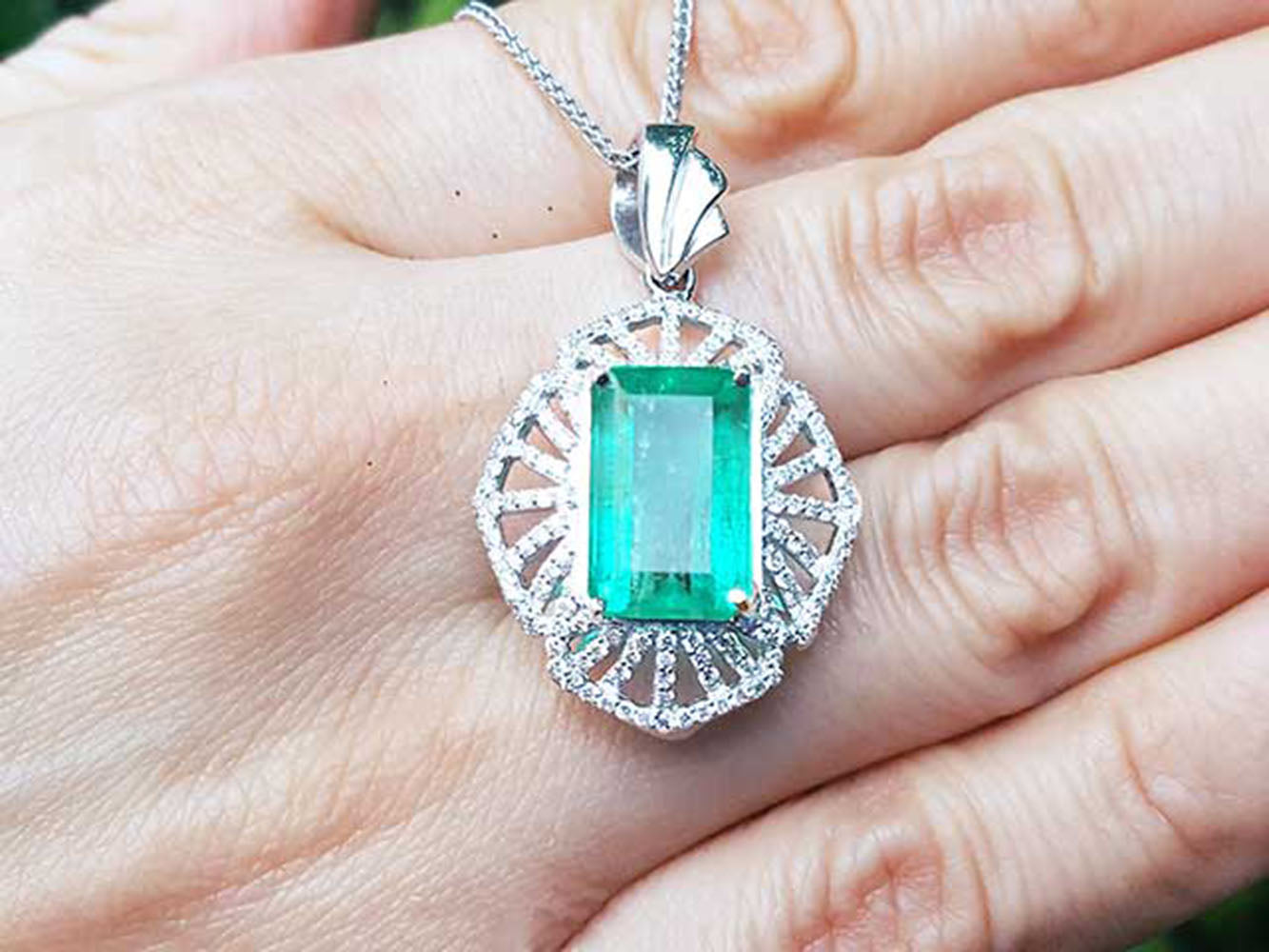 On a hand being displayed a natural emerald pendant with accent diamonds crafted in white gold.