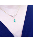 A close-up of a 14K yellow gold necklace with a baguette-cut Colombian emerald dangling from a diamond-studded V-shaped pendant, worn on a model's neck.