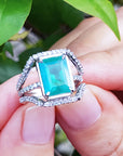 An emeral-cut-emerald ring with diamond accents held between fingers against a natural background.