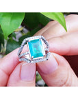 An emeral-cut-emerald ring with diamond accents held between fingers against a natural background.