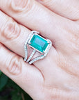 Ring with an emerald-cut emerald ring on a hand against a blurred natural background.