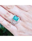 Ring with an emerald-cut emerald ring on a hand against a blurred natural background.