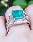 This is an emerald and diamond ring on a finger against a blurred green background.