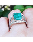 Emerald-cut 3.90 ct bluish-green emerald ring with triple diamond rows in 14K white gold on a finger.