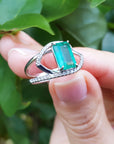 A rectangular emerald on a split shank ring held by a hand against a leafy background.