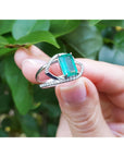 A rectangular emerald on a split shank ring held by a hand against a leafy background.