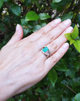 Hand wearing a white gold ring with a rectangular emerald against a green leafy background.