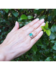 Hand wearing a white gold ring with a rectangular emerald against a green leafy background.