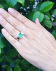 Hand wearing a gold ring with a natural emerald against a green leafy background.