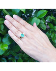 Hand wearing a gold ring with a natural emerald against a green leafy background.