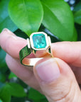 A colombian emerald solitaire ring held by a hand against a green leafy background.