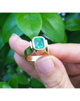 A colombian emerald solitaire ring held by a hand against a green leafy background.