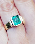 A women's solitaire emerald ring bezel set in yellow gold on a finger.