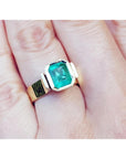 A women's solitaire emerald ring bezel set in yellow gold on a finger.