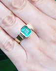 A bezel set emerald solitaire ring on a person's hand against a green leafy background.