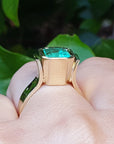 Viewing the underside of a natural emerald solitaire ring on a finger against a green leafy background.