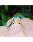 Viewing the underside of a natural emerald solitaire ring on a finger against a green leafy background.