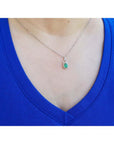Pear-shaped Colombian emerald and diamond halo pendant on a delicate 14K white gold chain, worn on a blue shirt.
